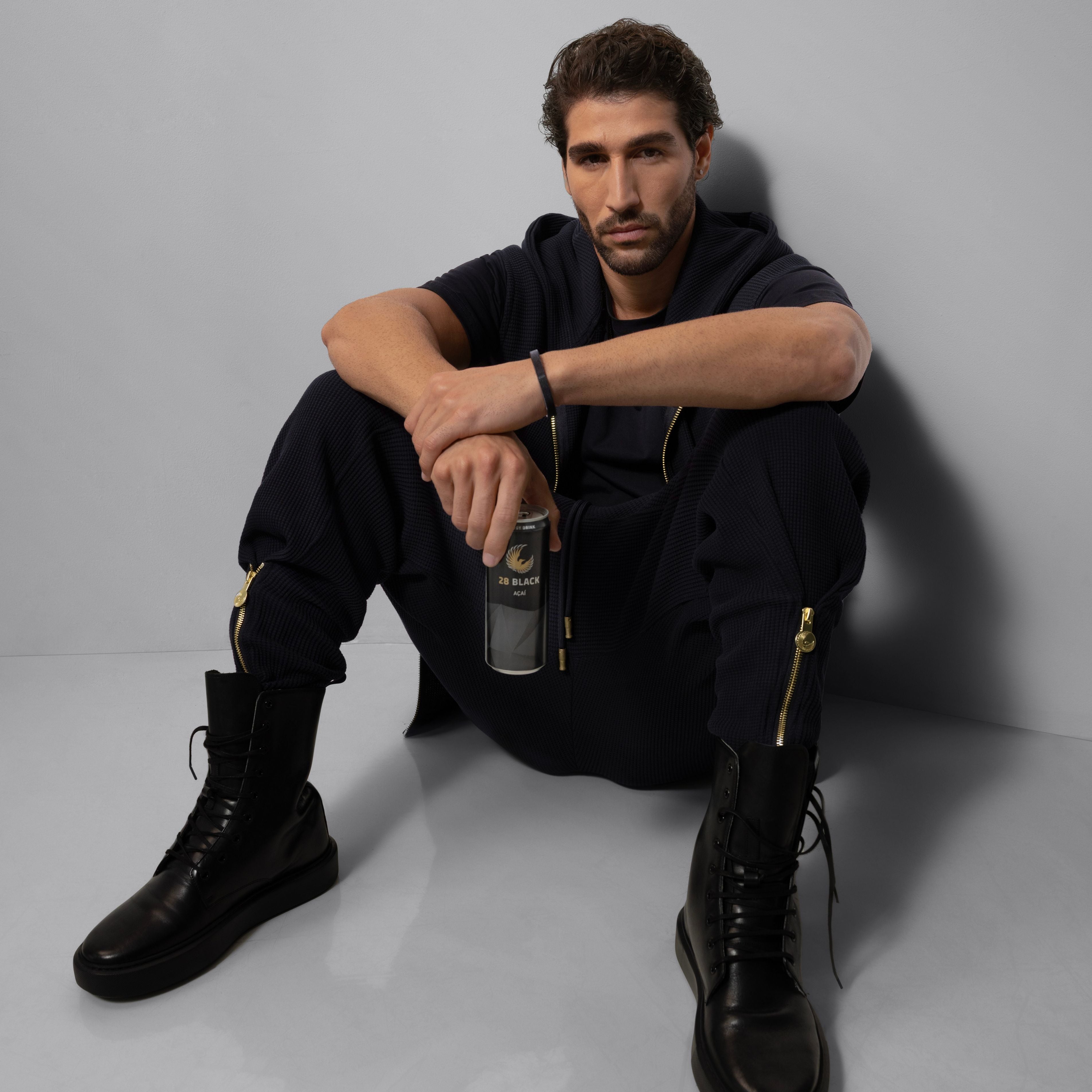 Man sitting on the floor wearing black boots and 28 BLACK outfit, holding a 28 BLACK energy drink can.